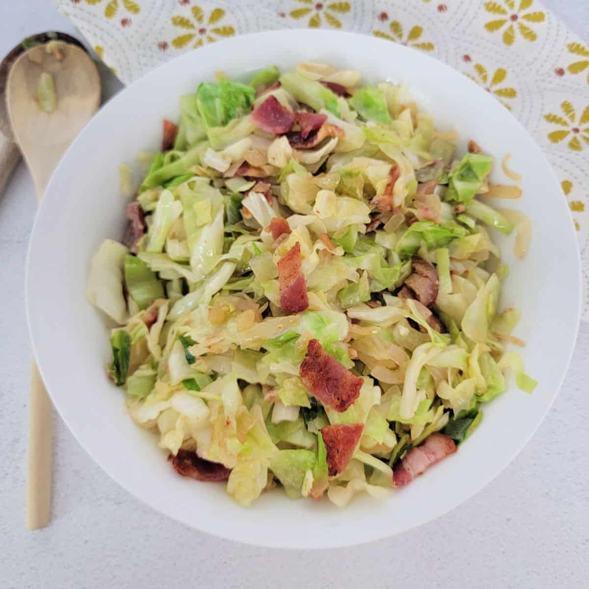southern fried cabbage with bacon in a white bowl next to a wooden spoon
