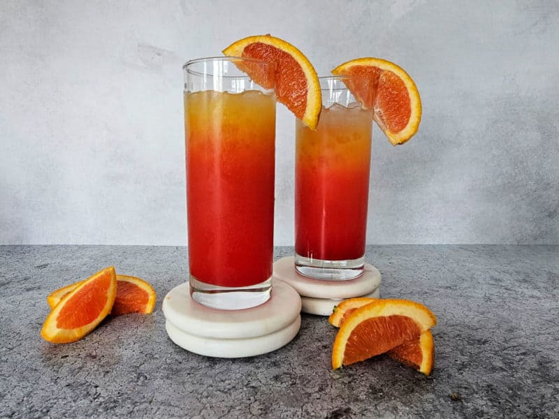 Two Vodka Sunrise drinks in tall glasses with orange wedge garnish on white coasters