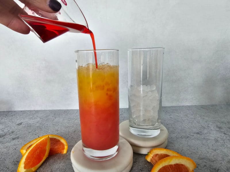 Grenadine pouring over orange juice in a tall glass with ice