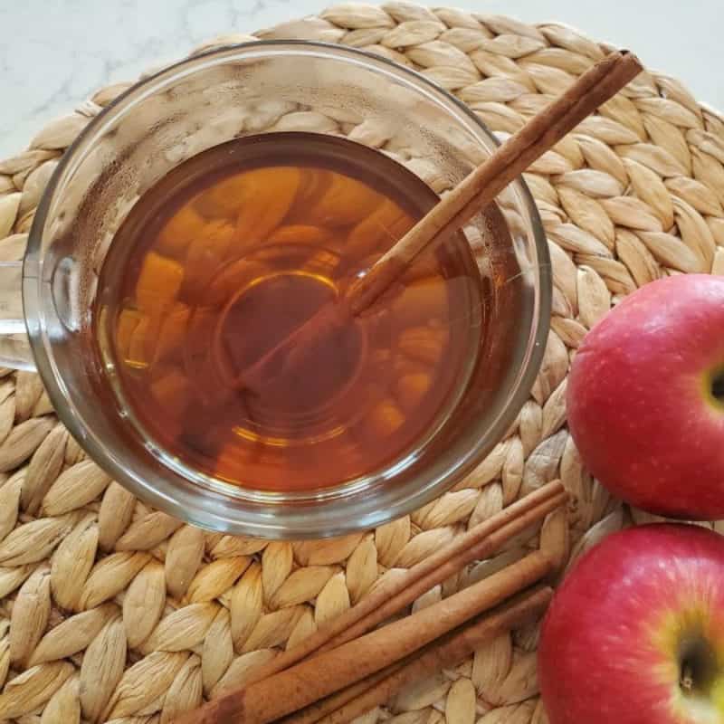 Caramel apple cider in a glass mug next to apples and cinnamon sticks