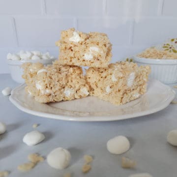 Classic rice krispie treats on a white plate surrounded by mini marshmallows and a bowl of rice krispies