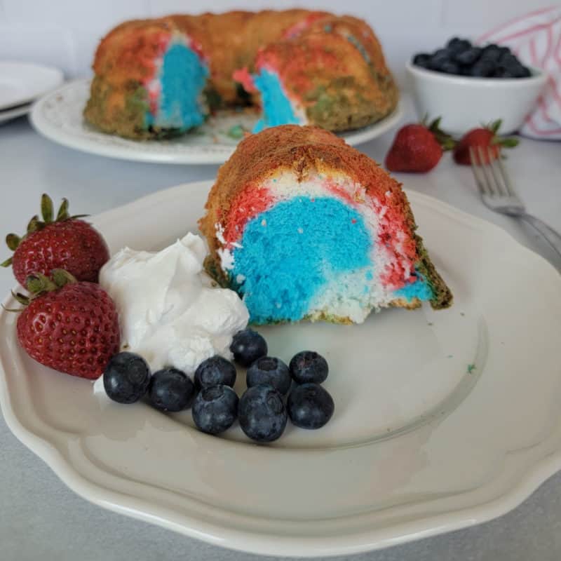 Red white and blue angel food cake next to strawberries, blueberries, and whipped cream