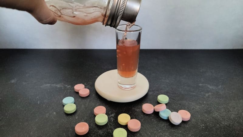 Cocktail shaker pouring pink liquid into a shot glass surrounded by sweet tart candies