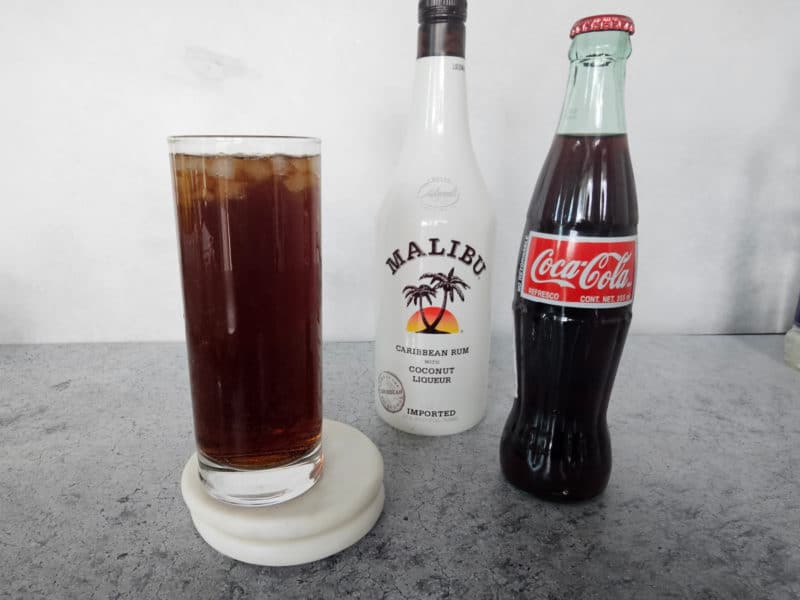 Tall glass filled with soda and ice next to bottles of Malibu Rum and a glass bottle of Coke