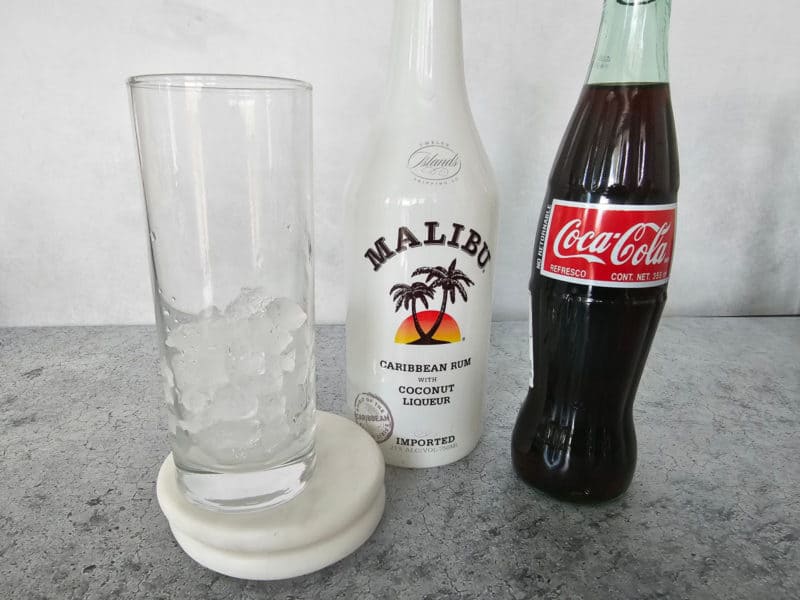 Tall grass with ice cubes, a bottle of Malibu Rum and a glass bottle of Coca Cola