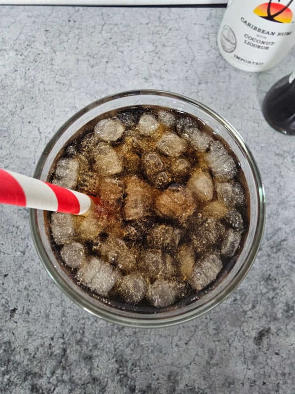 Looking down into a glass with ice and a red striped straw. Bottle of Malibu and edge of a bottle of coke in the background