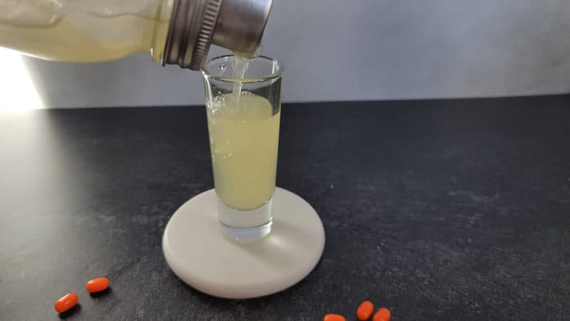 cocktail shaker pouring yellow liquid in a shot glass with tic tacs next to it