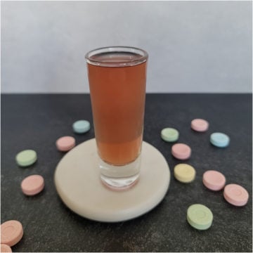 Sweet tart shot in a glass shot glass on a white coaster surrounded by Sweet Tart Candies
