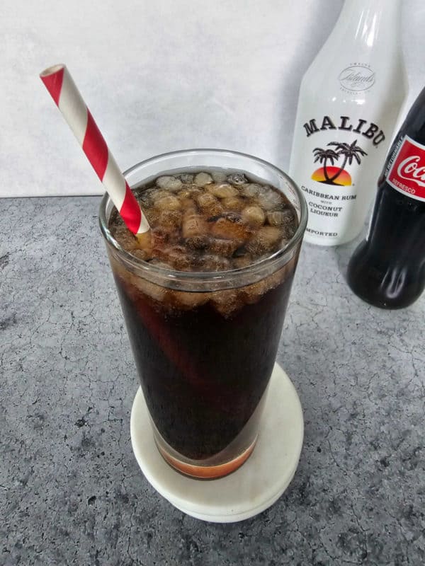 Tall glass with soda in it with a red striped straw near a bottle of Malibu Rum and Coca Cola