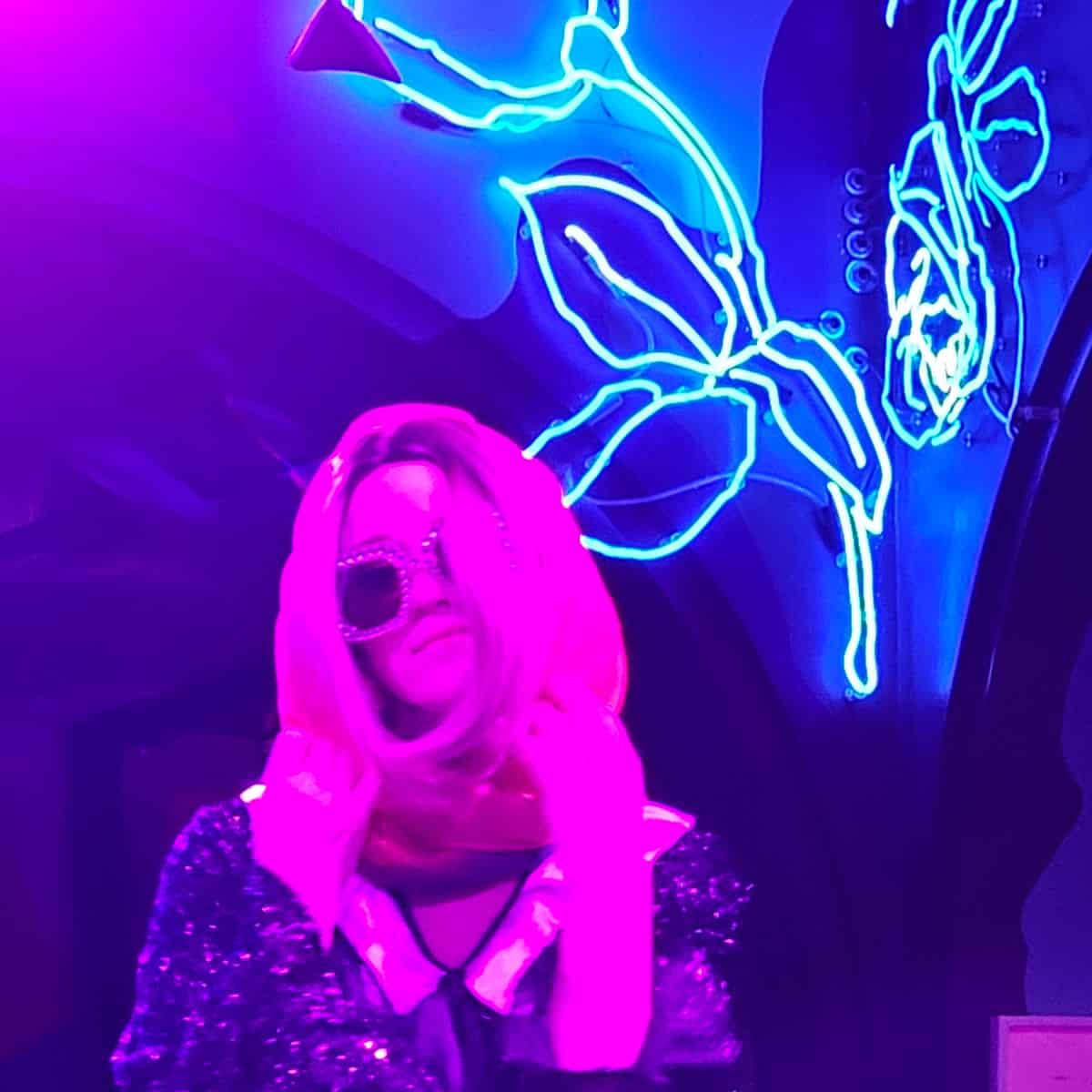 Woman with large sunglasses and a scarf next to a neon light at SuperFrico