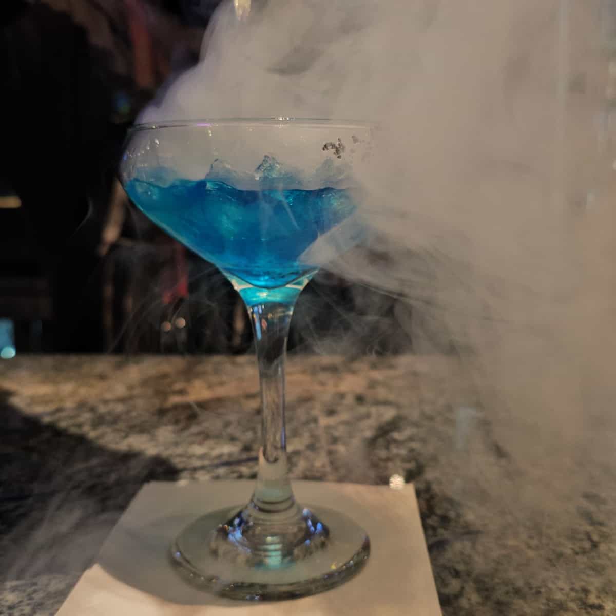 Blue cocktail with smoke coming out of it in a martini glass on a dark bar. 