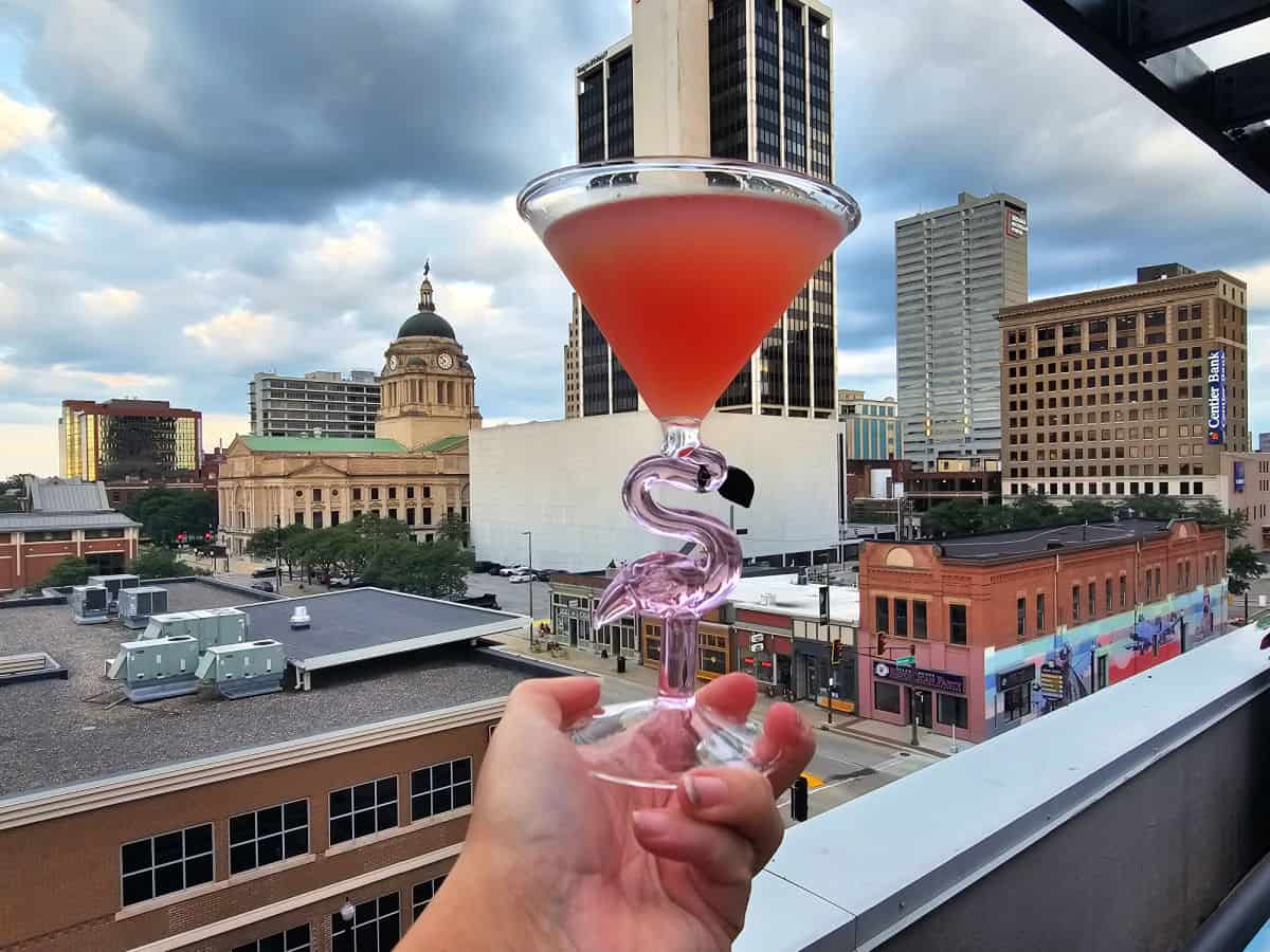Hand holding a pink drink in a flamingo glass with the Fort Wayne skyline in the background
