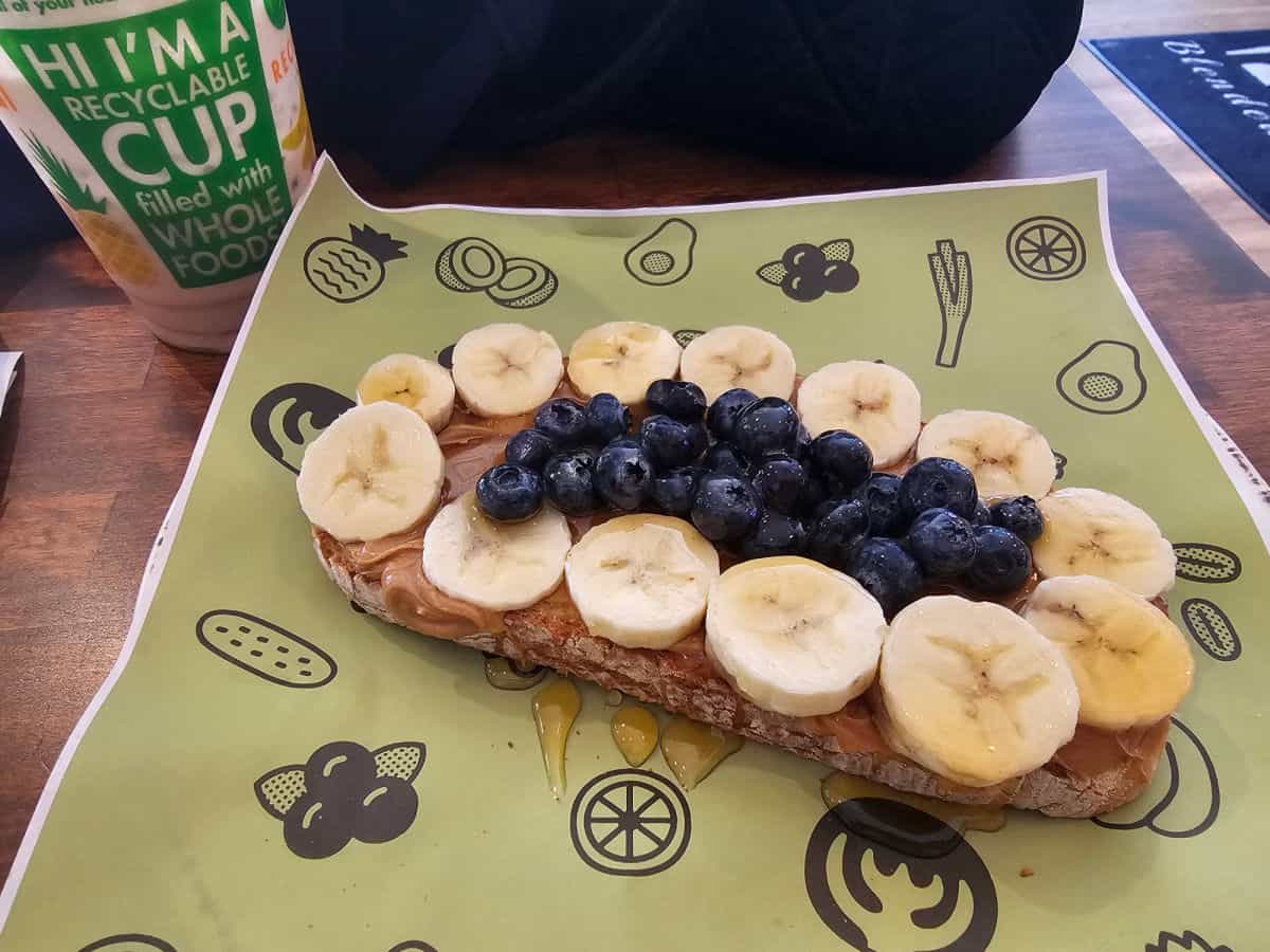 Toast with peanut butter, bananas, and blueberries on a green paper with Kanela on it next to a recylable coffee cup