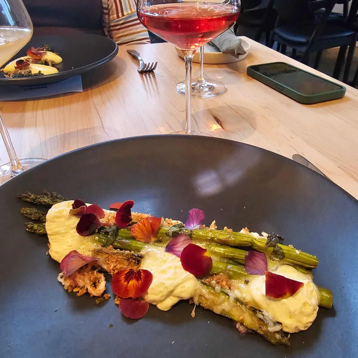 Asparagus on a dark plate with botanicals and cheese next to a rose wine