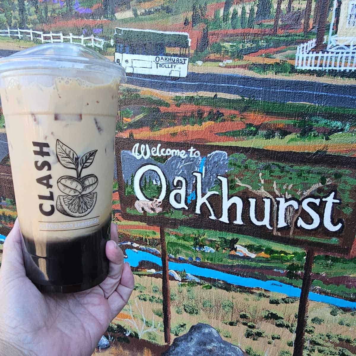 Hand holding a Class Coffee Cup in front of the Oakhurst mural