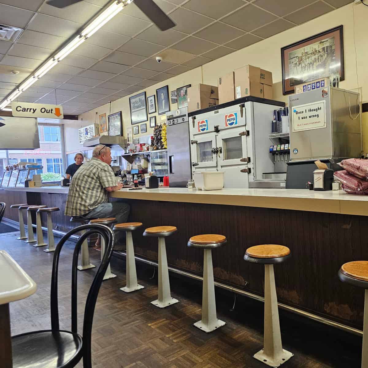 counter seating in Coney Island Hot Dog Fort Wayne with a pepsi sign, guy sitting at the counter, and carry out sign hanging from the ceiling