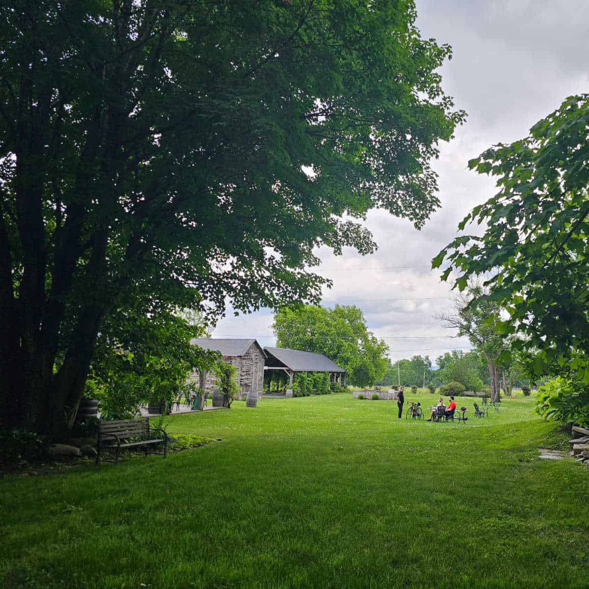 Green fields leading to a farmhouse and covered pavillion, people sitting at tables with bikes and wine