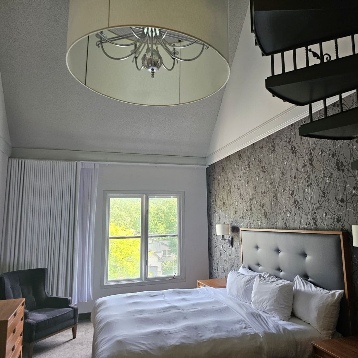 King bed with pillows next to a grey wallpapered wall, windows looking out to trees, large chandelier and chair