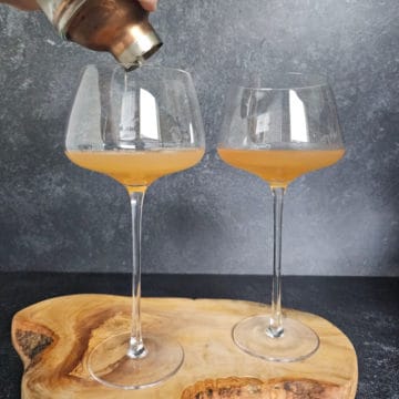 copper lidded cocktail shaker pouring into two tall coupe glasses with Hennessey Sidecar Cocktail in them sitting on a wooden board