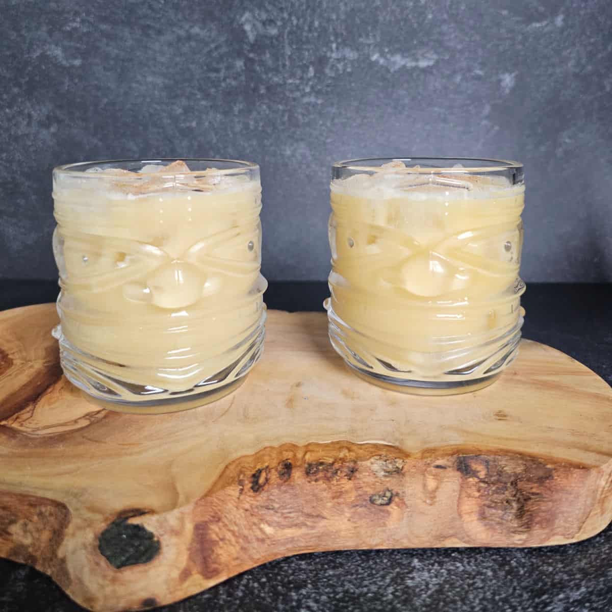 two tiki glasses with Painkiller Cocktails garnished with nutmeg sitting on a wooden board